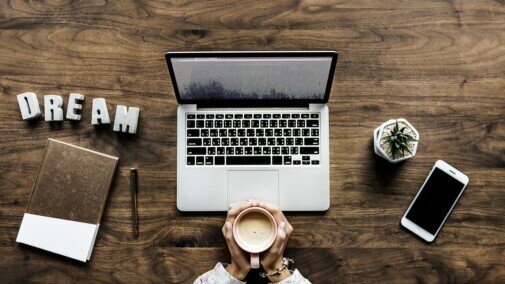 Open laptop, cellphone, small plant, person is griping their coffee cup with both hands, the word Dream in white block letters